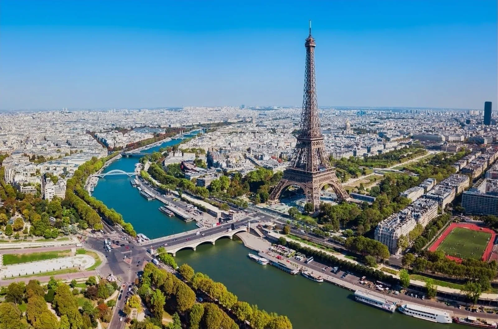 Vue aérienne de Paris en plein jour : la Tour Eiffel au centre, la Seine et ses ponts serpentant entre les toits, jusqu'à l'horizon dégagé