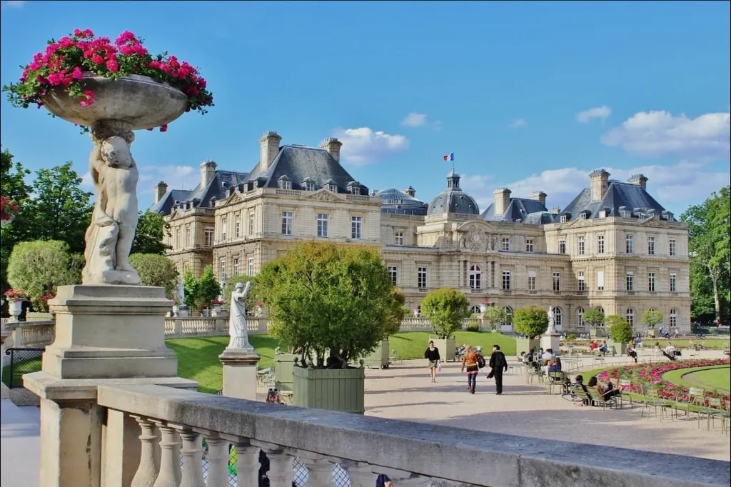 Terrasse du jardin du Luxembourg en plein jour : vasque fleurie portée par des statues au premier plan, palais du Luxembourg et promeneurs au fond